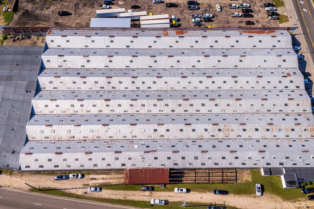 An aerial view of a long commercial warehouse with a low-slope metal roof beside a road and a parking lot