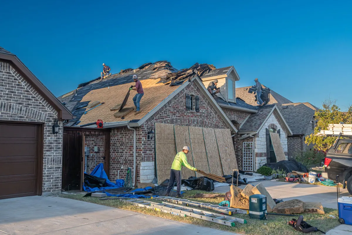 A roofing crew tearing off old shingles down to the decking on a brick house, one roofer carrying sheathing across the lawn below